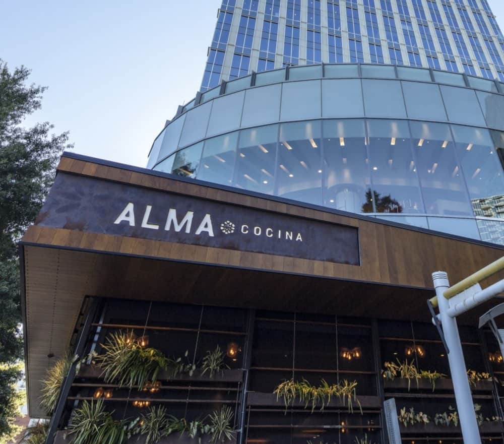 Exterior view of a modern building with large glass windows and a sign reading "ALMA COCINA" above the entrance, with greenery and trees visible.