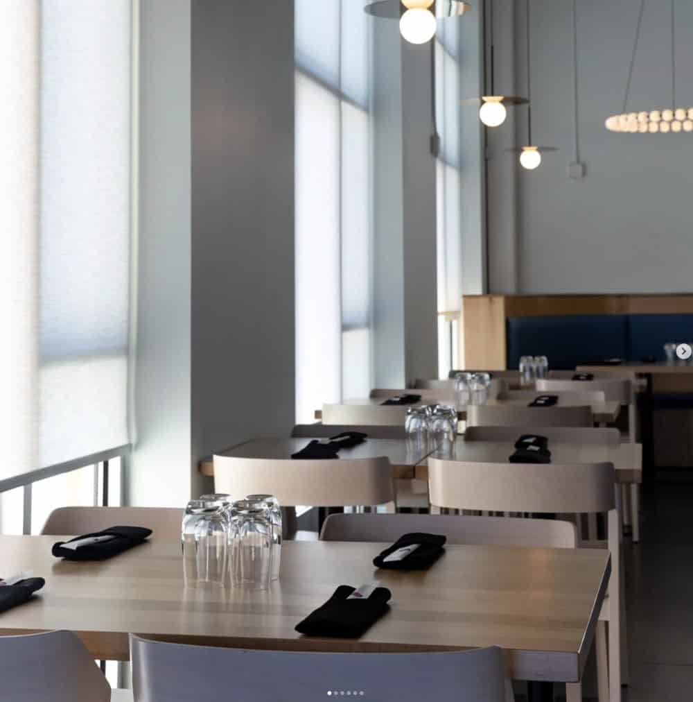 A modern, minimalistic restaurant interior with wooden tables set with black napkins and stacked glasses. Hanging pendant lights illuminate the space.