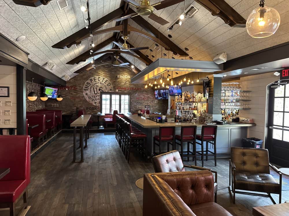 A modern bar with high ceilings features a long bar counter, red booth seating, wooden beams, and flat-screen TVs. The space has a mix of rustic and contemporary decor elements.