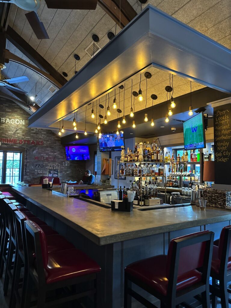 A modern bar with a concrete countertop, red cushioned bar stools, and hanging Edison bulb lights. Shelves behind the bar are stocked with various bottles, and televisions display sports games.