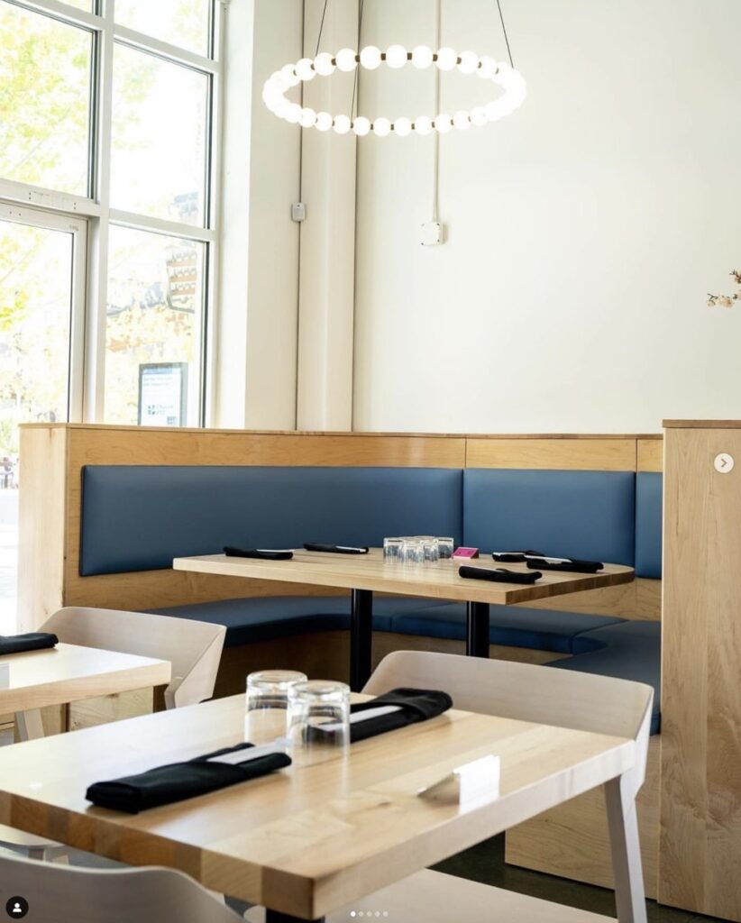 A modern dining area with wooden tables, blue cushioned booth seating, and black napkins. A circular chandelier with bright bulbs hangs from the ceiling. Large windows bring in natural light.