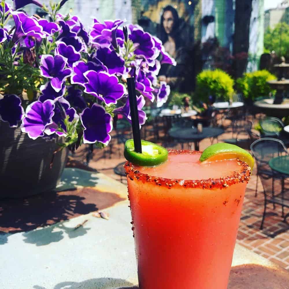 A red cocktail in a glass with a salted rim, garnished with lime and jalapeño slices, sits on a table near purple flowers in an outdoor patio setting.