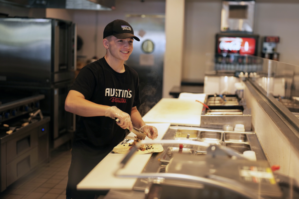 Person in a kitchen smiling, wearing a cap and "Austin's Grill" shirt, using a spatula to prepare food on a counter with kitchen equipment and soda dispenser in the background.