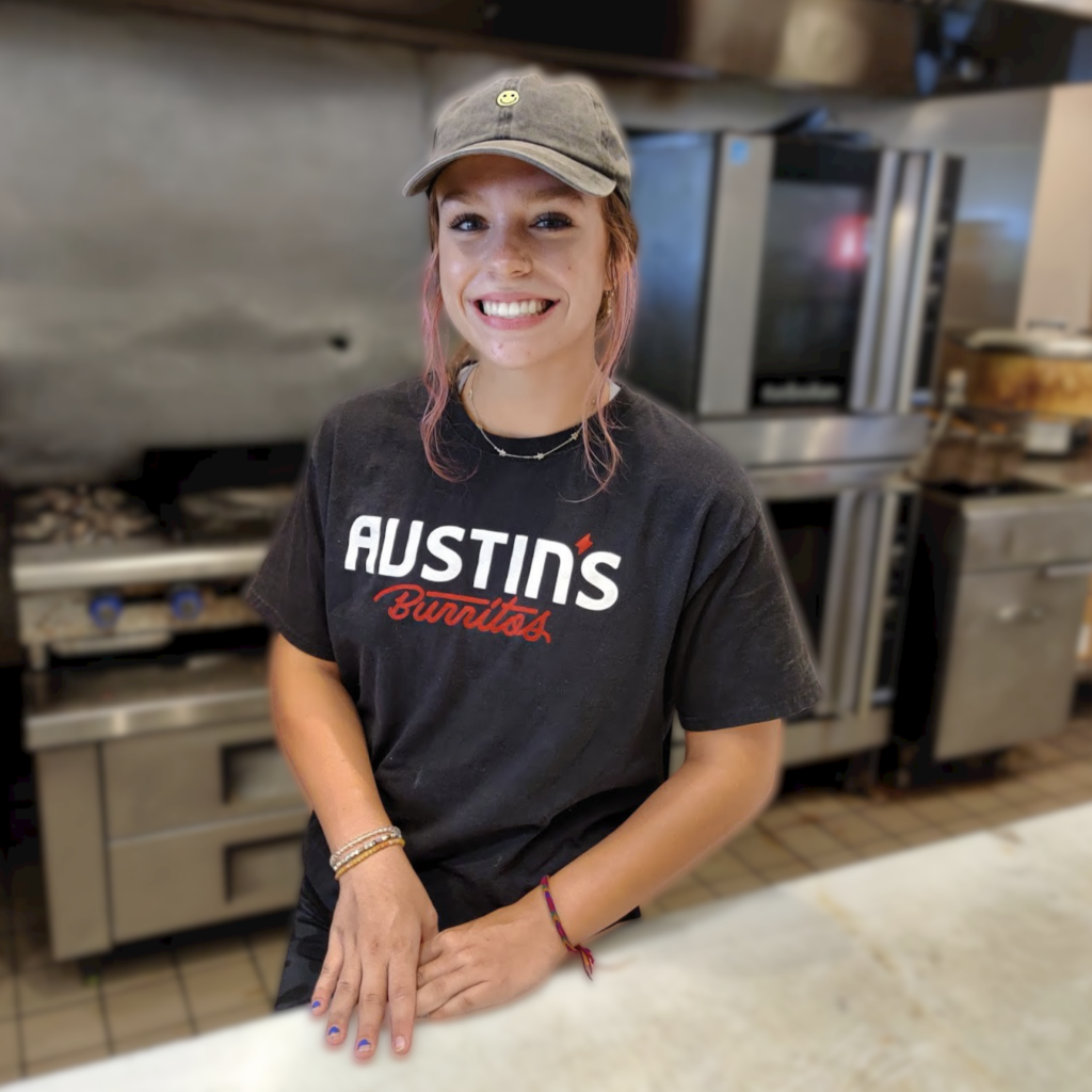 Person wearing a "Austins Burritos" t-shirt and cap smiles while standing in a commercial kitchen.