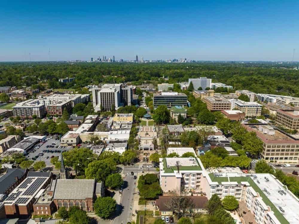 Aerial view of a city neighborhood with mid-rise buildings, tree-lined streets, and a distant skyline under a clear blue sky.