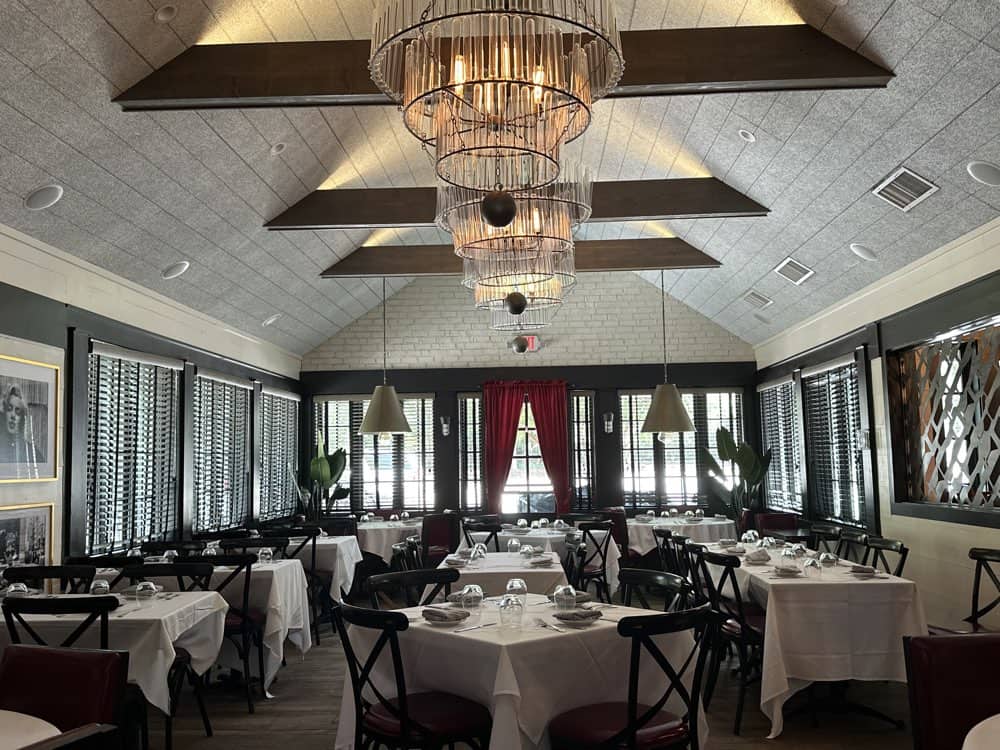 An elegant dining room with neatly arranged tables covered in white tablecloths, black chairs, and modern chandeliers hanging from the ceiling. Red curtains and green plants provide accents.