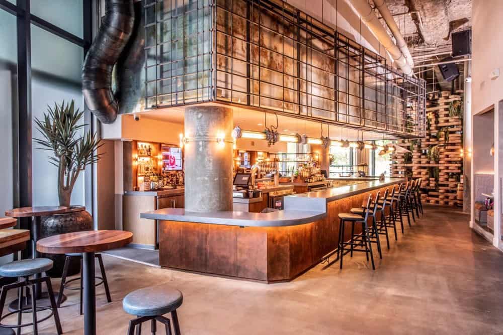 Modern bar interior with high ceilings, metal and wood accents, a long counter with barstools, ambient lighting, and shelves filled with stacked logs in the background.