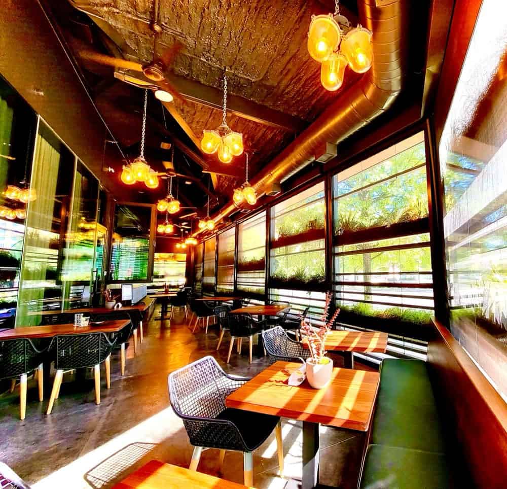 A modern restaurant interior with wooden tables, black chairs, hanging industrial lights, and large windows letting in bright sunlight.