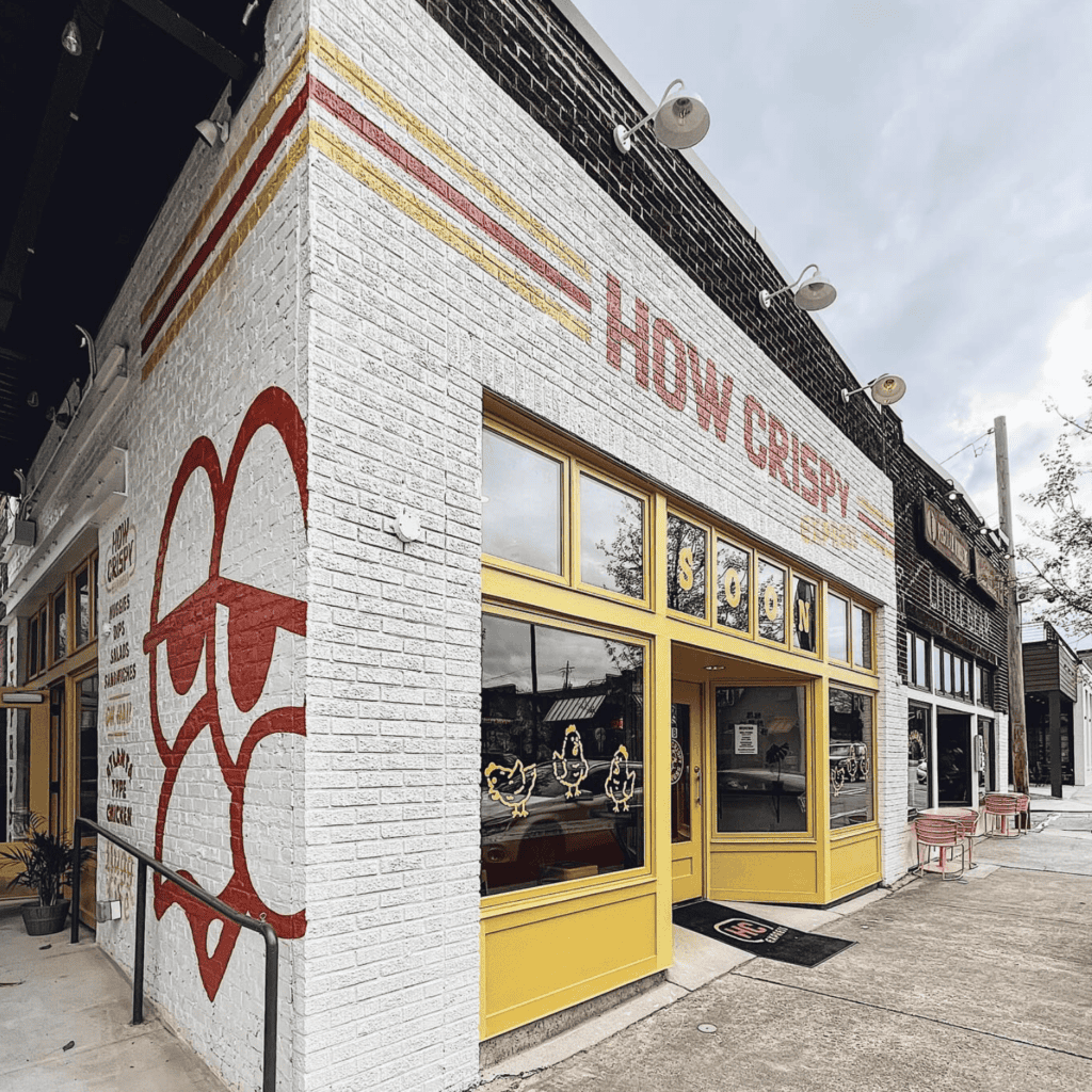 A white brick building with yellow-framed windows and a mural reading "HOW CREEPY" on the wall, featuring red and yellow decorative designs.