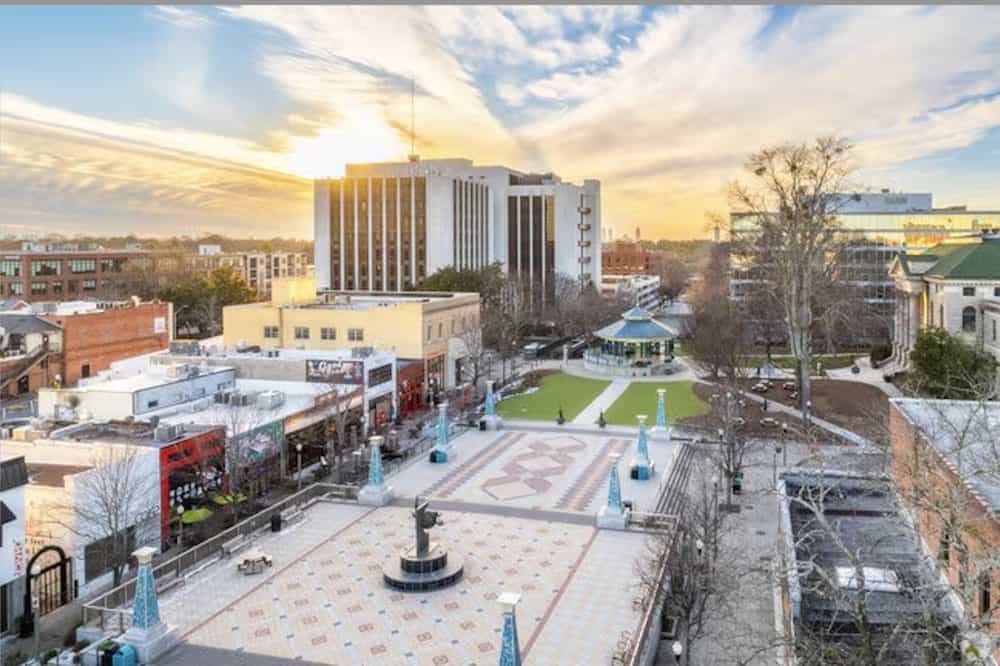 A city plaza with patterned paving, a central fountain, surrounding buildings, a park area, and a tall office building under a partly cloudy sky at sunset.
