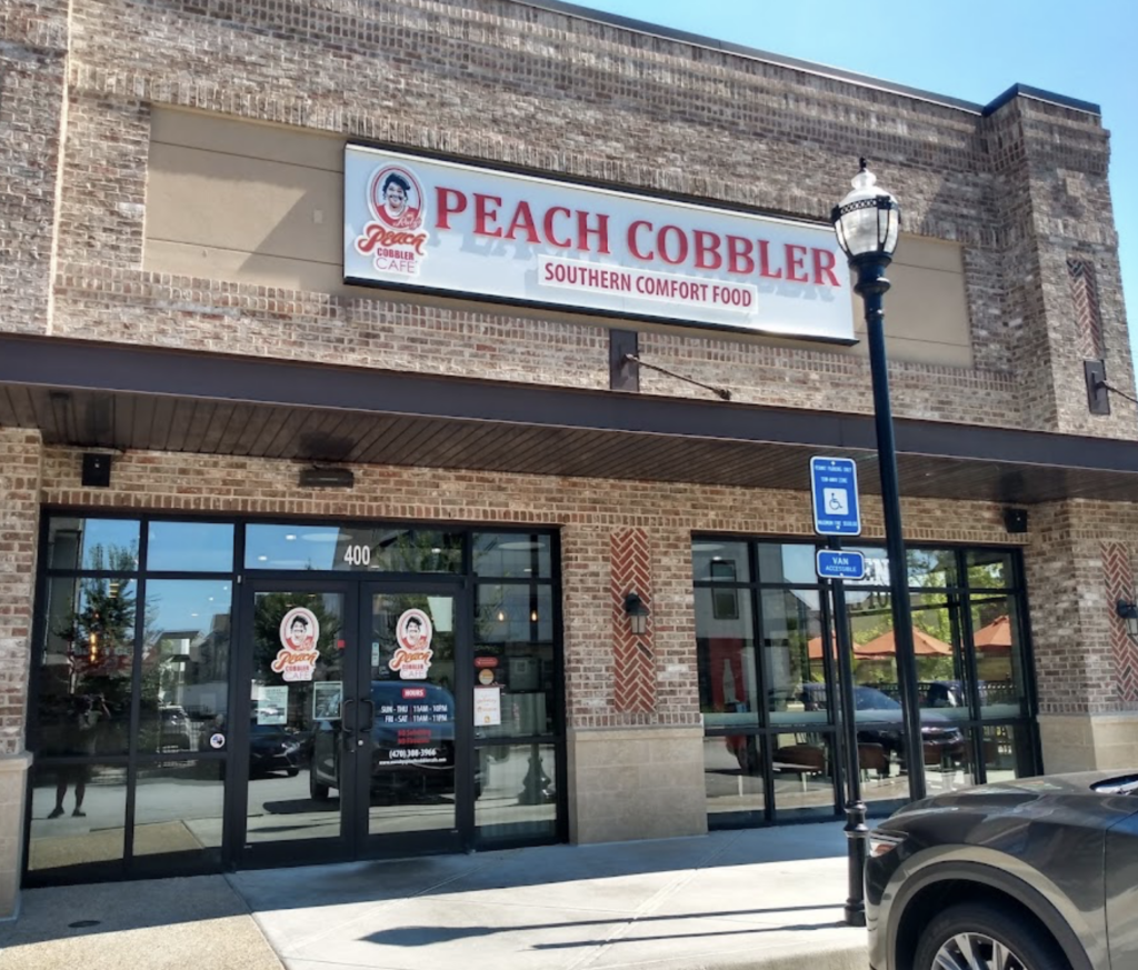 Brick building with signage reading "Ms. Ruby's Peach Cobbler Café: Southern Comfort Food." Glass entrance doors and a handicap parking sign are visible, capturing the essence of Southern charm in Smyrna, GA.
