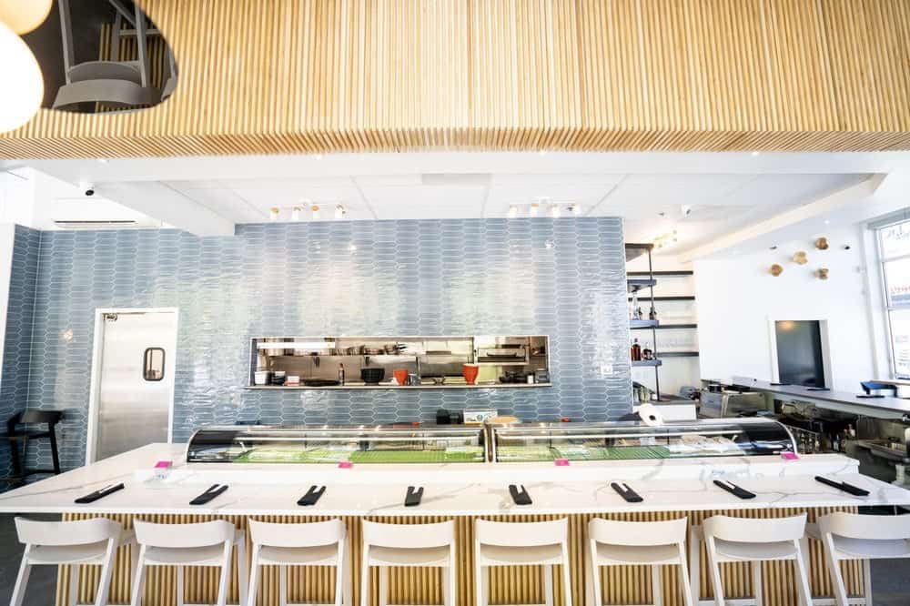 Modern sushi restaurant interior with a long bar counter lined with white stools, and a tiled wall behind displaying an open kitchen.