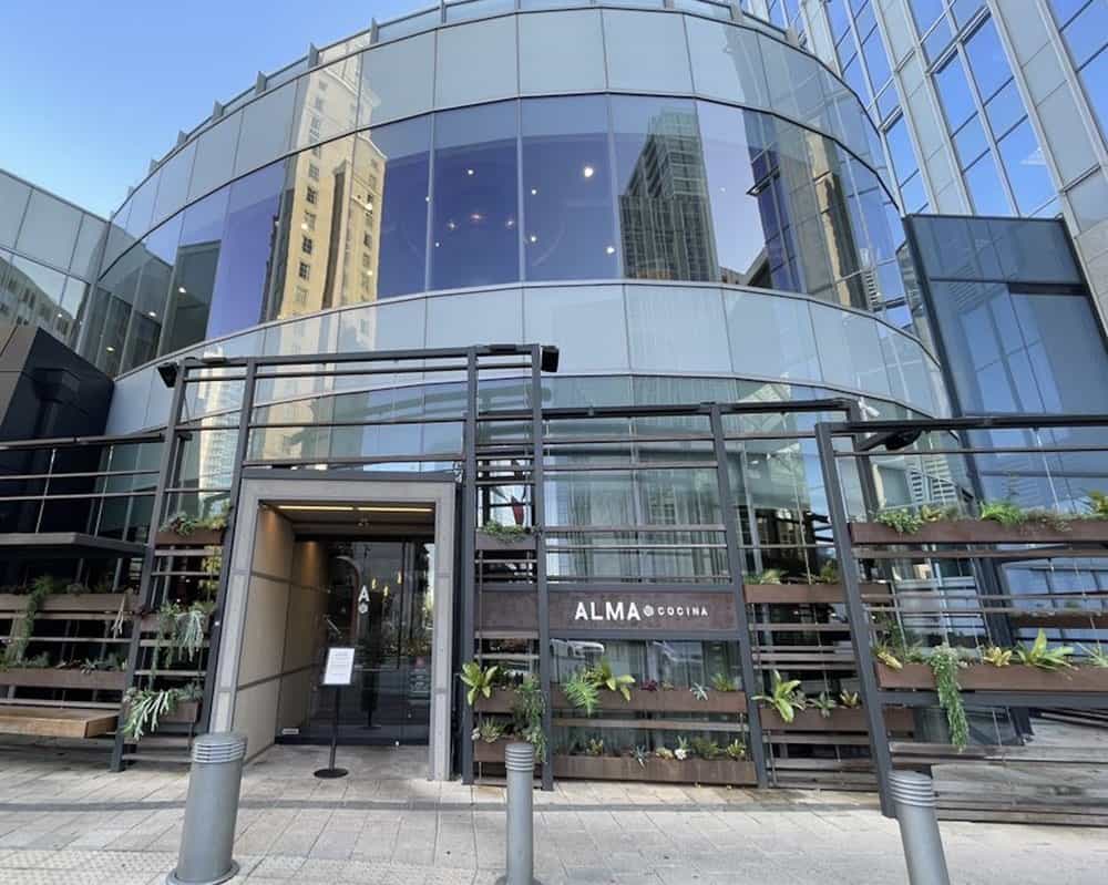 Glass-fronted modern building with a curved facade, featuring the entrance to ALMA Cocina restaurant and vertical planters with greenery.