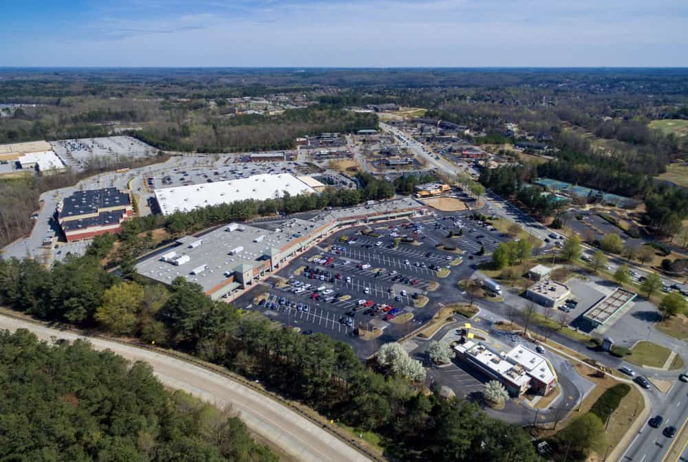 Aerial view of a suburban shopping center with multiple parking lots, surrounded by trees and roads. The scene includes several large buildings and scattered vehicles.