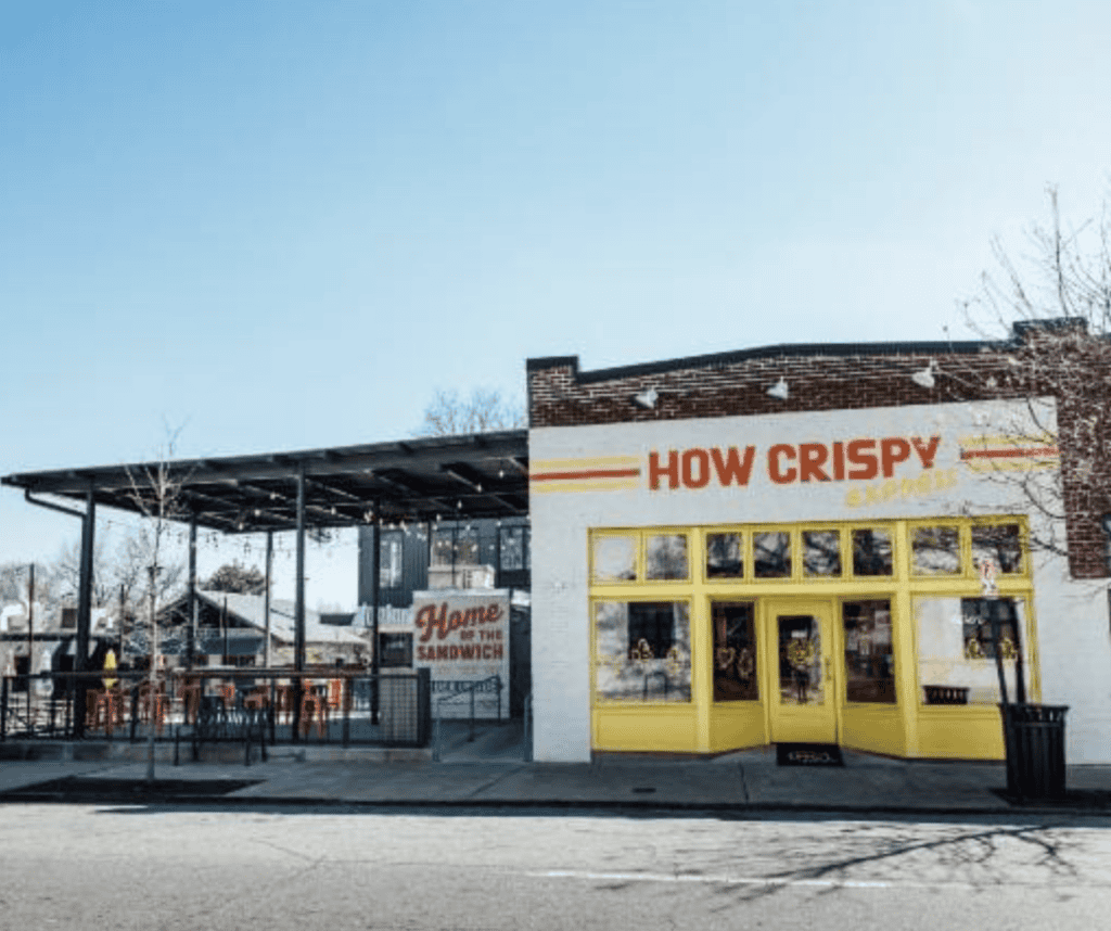 Exterior of How Crispy Express restaurant with yellow-trimmed windows, bold signage, and a covered outdoor patio with string lights and red bar stools.