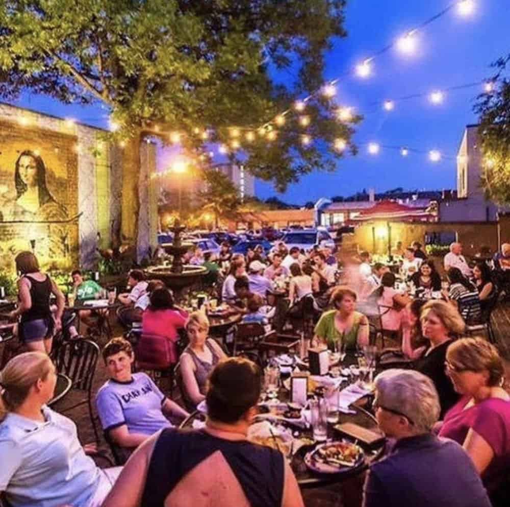 A busy outdoor restaurant patio at dusk with many people dining under string lights; a mural of the Mona Lisa is visible on a wall in the background.