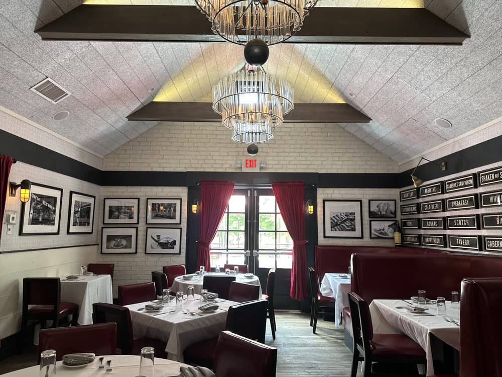 A restaurant interior with several tables covered in white tablecloths, red chairs, framed photos on the walls, and two ceiling chandeliers. Red curtains adorn the windows, letting in natural light.