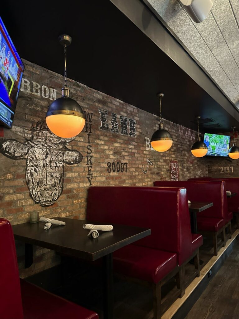 Image of a cozy restaurant interior with brick walls, red leather booths, hanging light fixtures, and TV screens. Artwork and text about beef and whiskey adorn the walls. Tables are set with napkin-wrapped utensils.