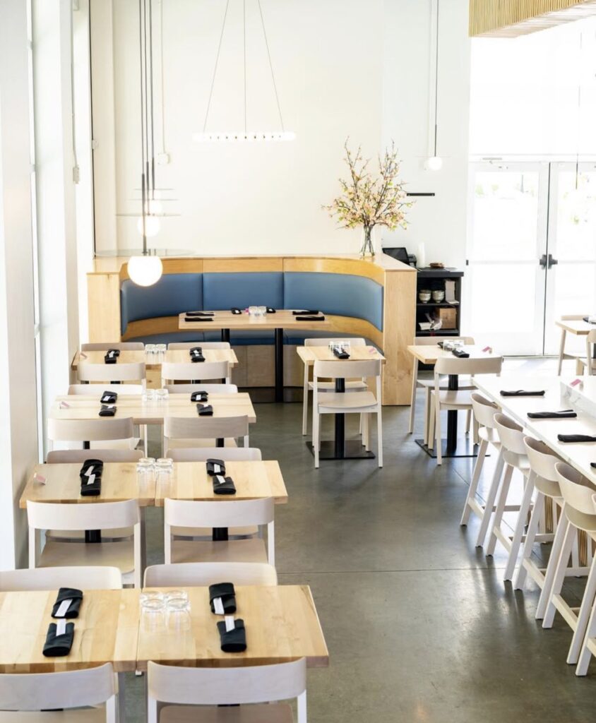 Modern, bright restaurant interior with wooden tables and chairs, set with napkins and utensils. A booth in the corner features a vase with flowers, and large windows provide natural light.