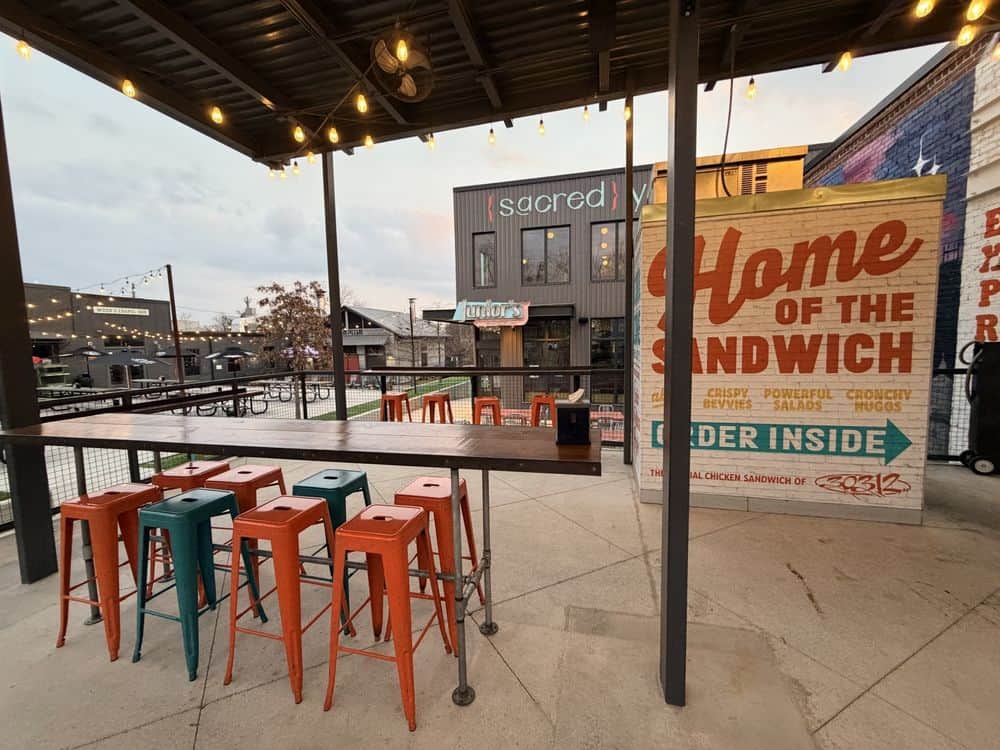 Outdoor patio with colorful metal stools, a long high table, string lights overhead, and a large sign reading "Home of the Sandwich" next to building entrances.