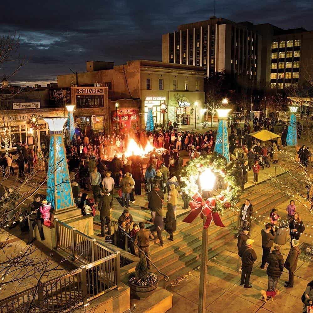 A crowd gathers around a fire pit in a decorated city plaza at night, with holiday lights and wreaths, and groups of people socializing on the steps and walkways.