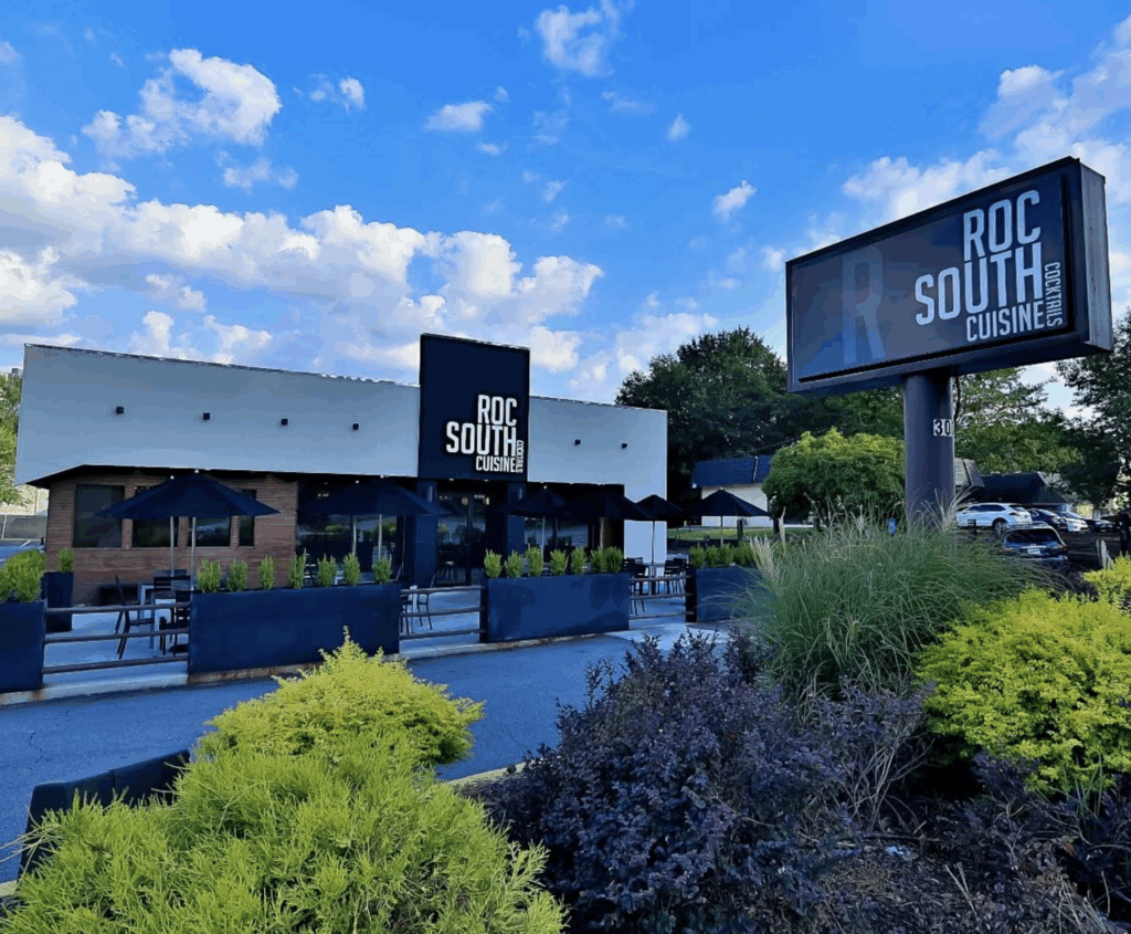 Outdoor view of ROC South Cuisine restaurant near Tribute Lofts, featuring patio seating with black umbrellas, prominent signage, lush greenery, and a partly cloudy sky—an inviting spot just steps from the O4W Beltline.