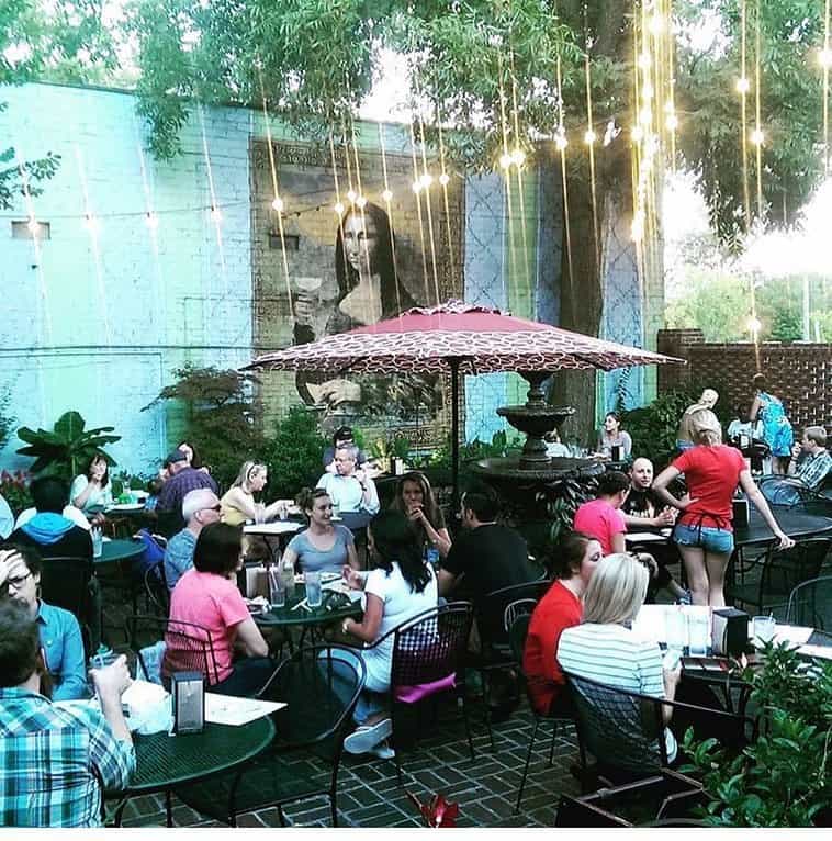Outdoor café with people dining at tables under string lights and umbrellas, featuring a large mural of the Mona Lisa on a blue wall in the background.