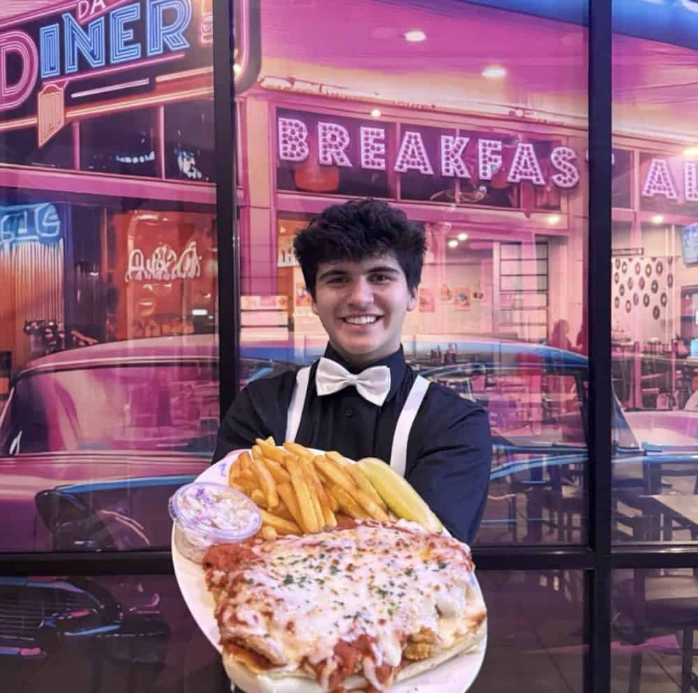 Young man holding pizza and fries outside Atlanta diner with neon signs.