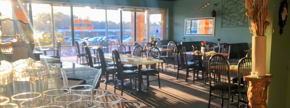 Sunlight streams into a Johns Creek Thai Restaurant's dining area with empty tables and chairs, glassware in the foreground, and a view of the parking lot through large windows.
