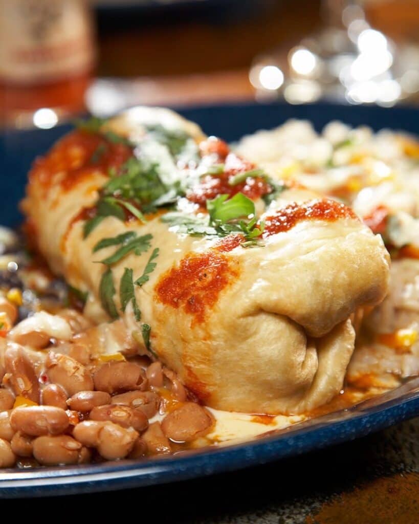 A close-up of a burrito topped with sauce and cilantro, served with pinto beans and rice on a blue plate.