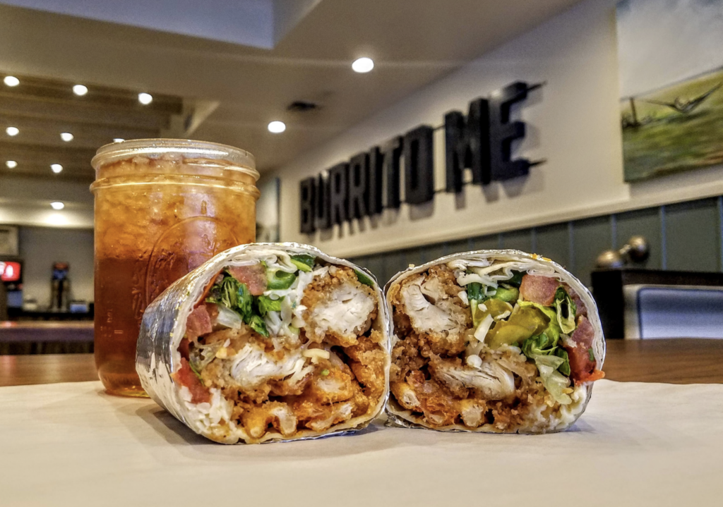 A burrito cut in half showing chicken, lettuce, tomatoes, and cheese, placed beside a glass of iced tea with a blurred "Burritofe" sign in the background.