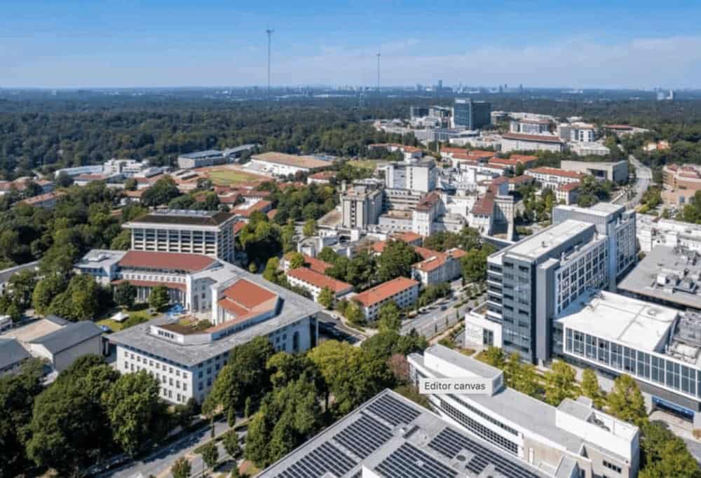 Aerial view of a large university campus with multiple academic buildings, green spaces, and surrounding trees under a clear sky.
