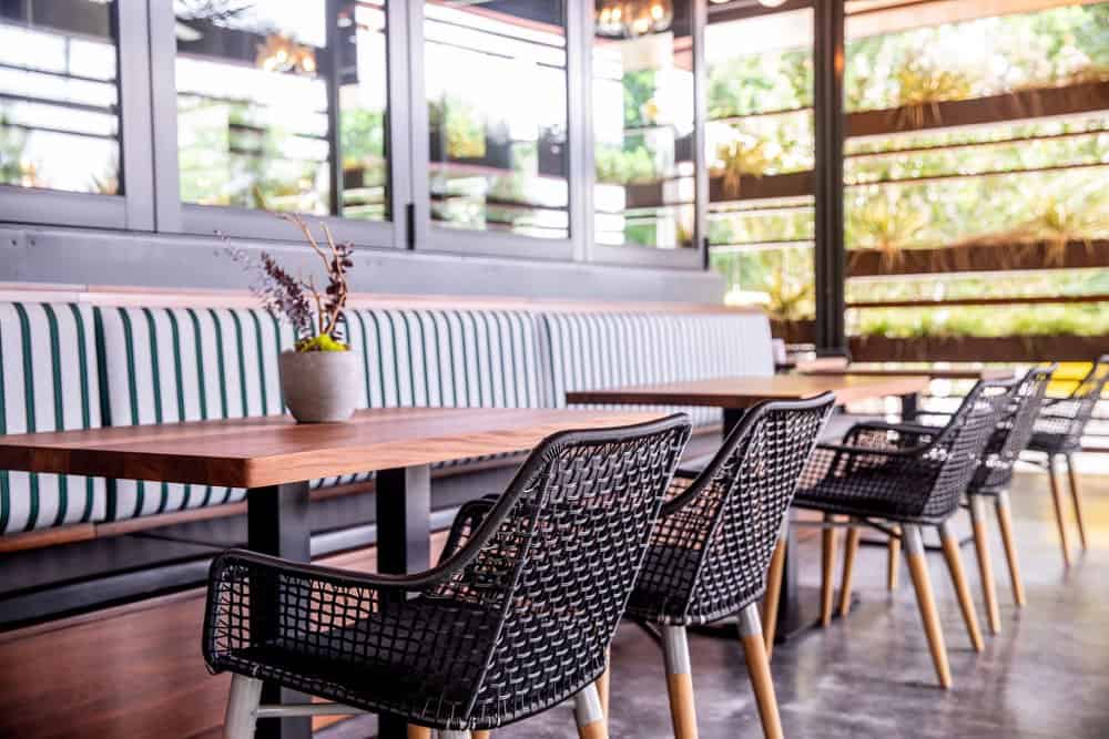 Modern cafe interior with wooden tables, striped cushioned bench seating, black woven chairs, and a small plant centerpiece, featuring large windows and natural light.