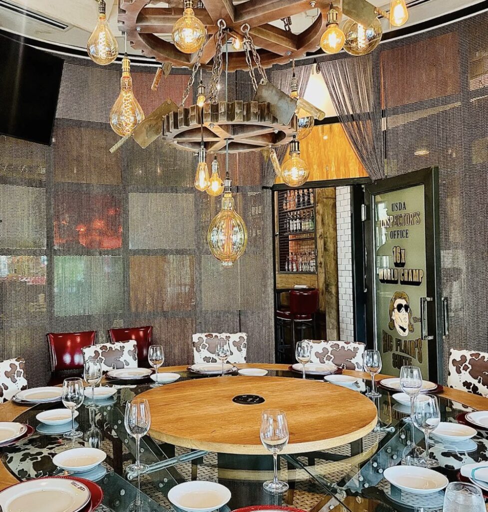 A dining room with a round table set for a meal, featuring unique hanging lights and cow-patterned chairs. A door in the background reads "USA Headmaster's Office, Big Plans 20k.