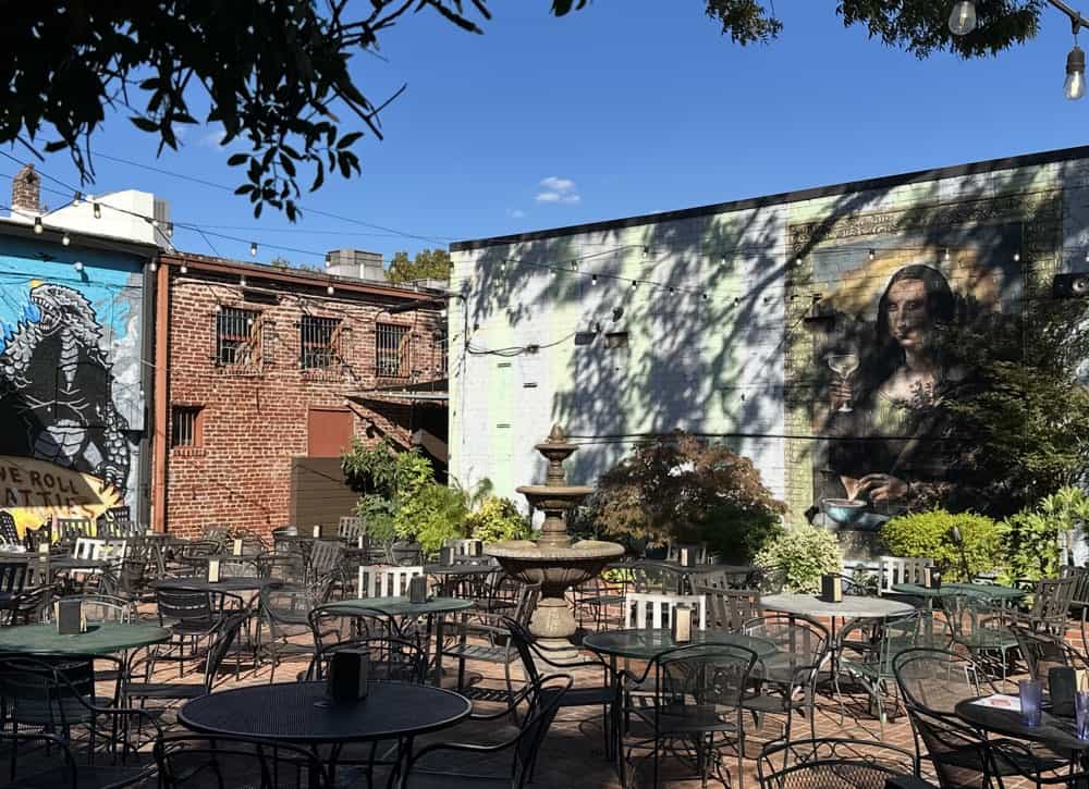 Outdoor patio with empty metal tables and chairs, a central fountain, and murals painted on surrounding brick and stucco walls under a clear blue sky—ideal setting for a Decatur GA restaurant in historic downtown Decatur.