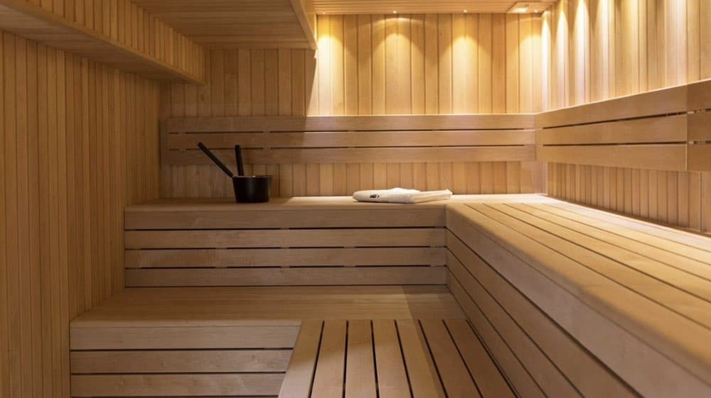 Wooden sauna interior with tiered benches, a bucket, and ladle. Soft lighting illuminates the wooden panels. A white towel is placed on one of the benches.