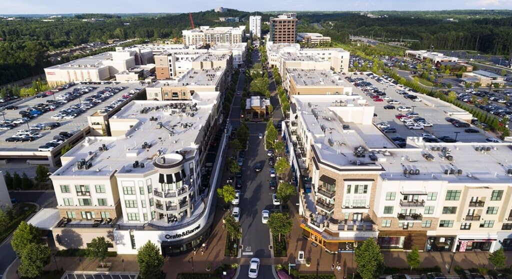 Aerial view of a modern mixed-use development with retail stores, apartments, tree-lined streets, and large parking lots, surrounded by greenery and distant buildings.
