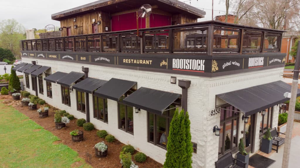 Two-story white brick building with black awnings and a balcony, featuring signs for ROOTSTOCK Restaurant and bar, located in vibrant Downtown Woodstock GA.