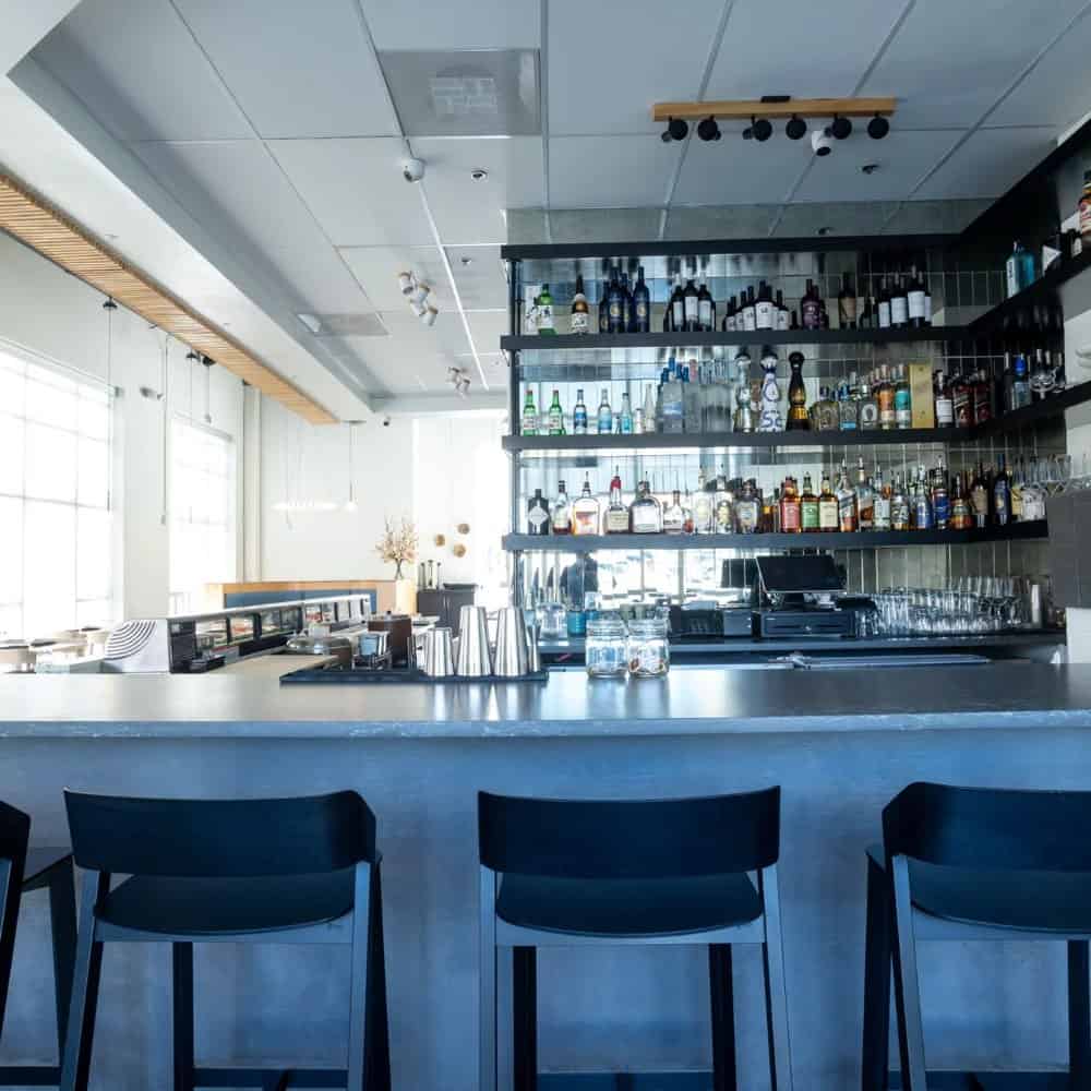 Modern bar interior with a sleek countertop, four black bar stools, and a fully stocked wall-mounted shelf with various bottles of alcohol. Natural light streams in through large windows.