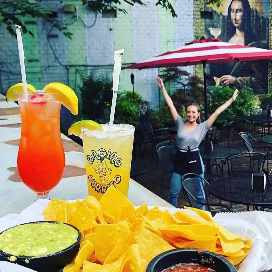 A table with nachos, guacamole, salsa, and two cocktails; a woman stands smiling with arms raised in an outdoor patio, with a Mona Lisa mural in the background.