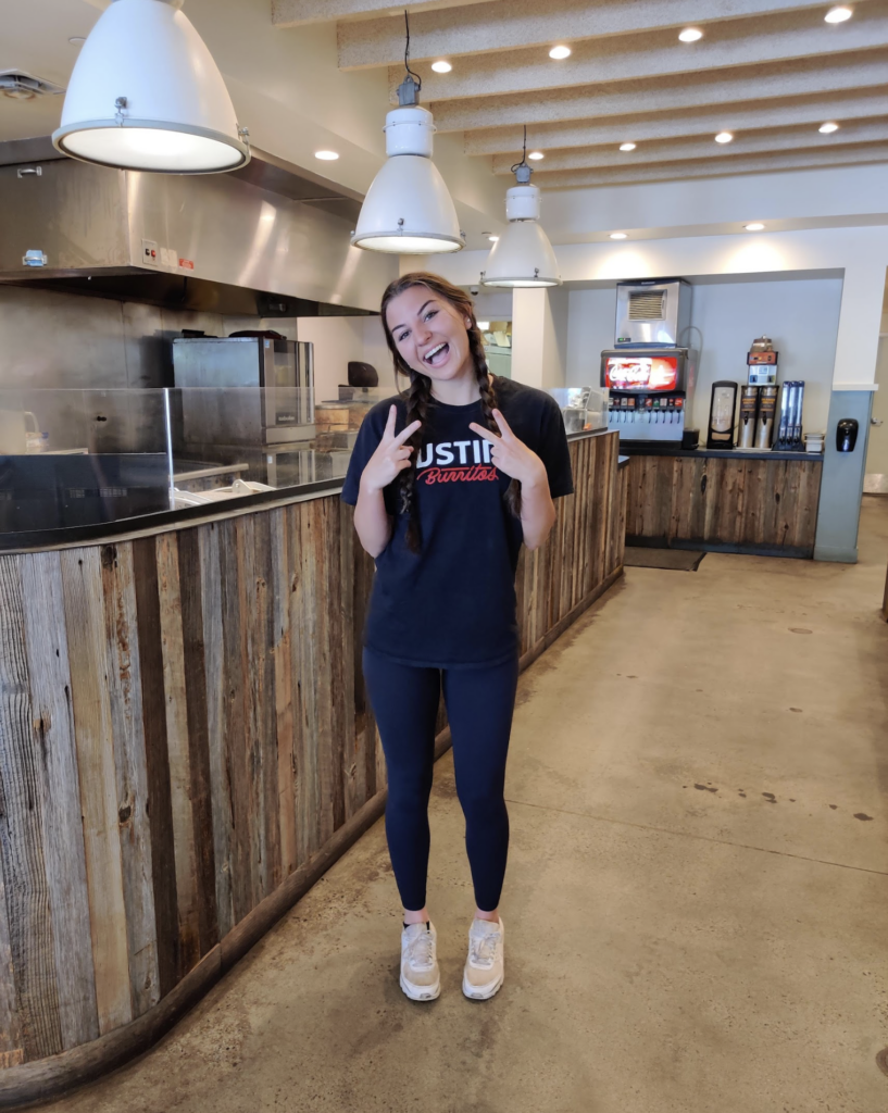 Person in casual attire stands indoors near a counter, smiling and gesturing with two peace signs.