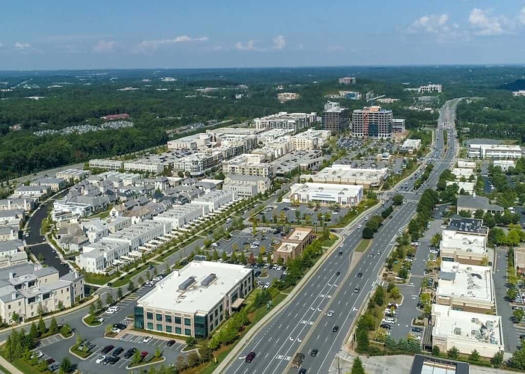 Aerial view of a suburban area with residential buildings, commercial properties, main roads, and surrounding greenery under a clear sky.