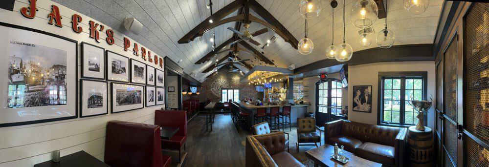 Panoramic view of a cozy, rustic-style cafe with leather seating, wooden beams, framed photos on the wall, an arched ceiling, and pendant lighting. The space includes a bar area and large windows.