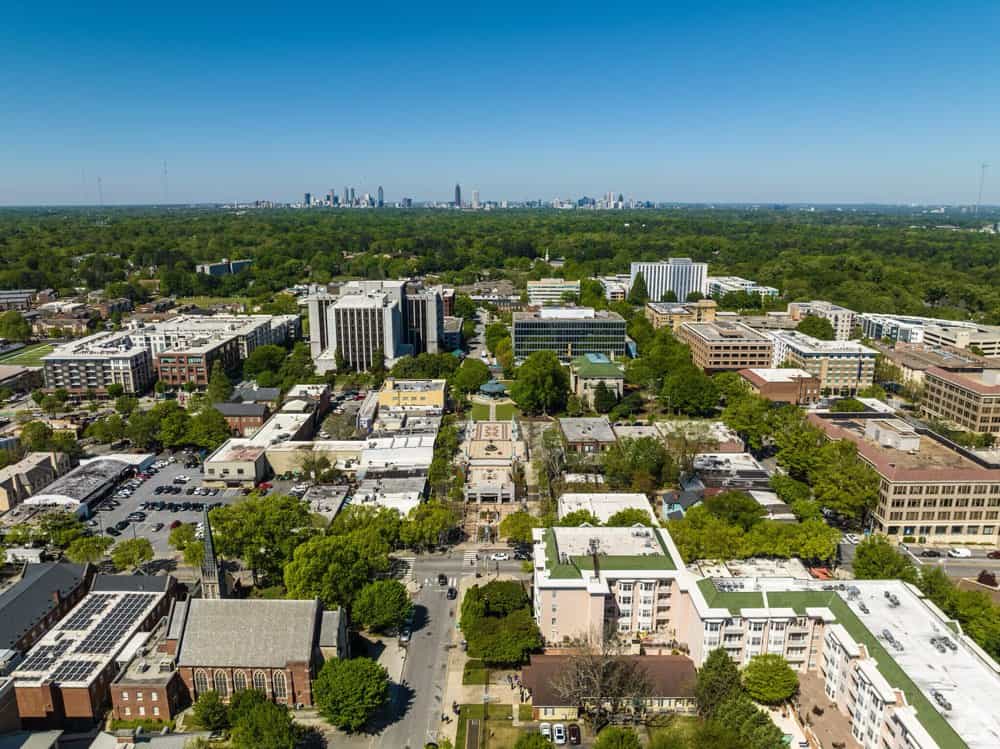 Aerial view of a suburban area with mid-rise buildings, tree-lined streets, and a distant city skyline under a clear blue sky.