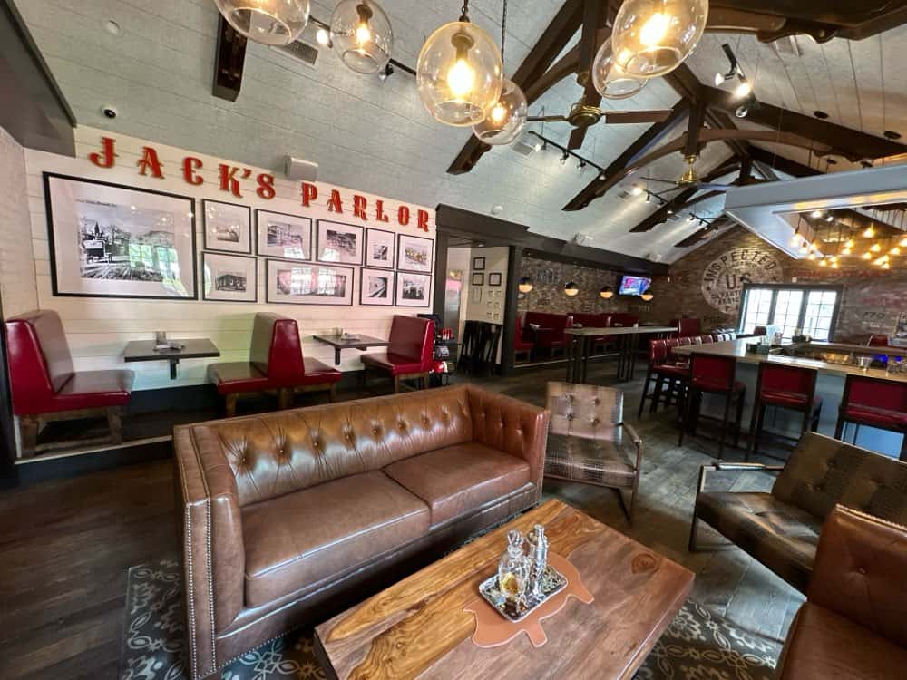 A cozy parlor featuring leather sofas, rustic tables, red booth seating, and a bar area. The walls showcase framed pictures and the lettering "Jack's Parlor" is displayed prominently.