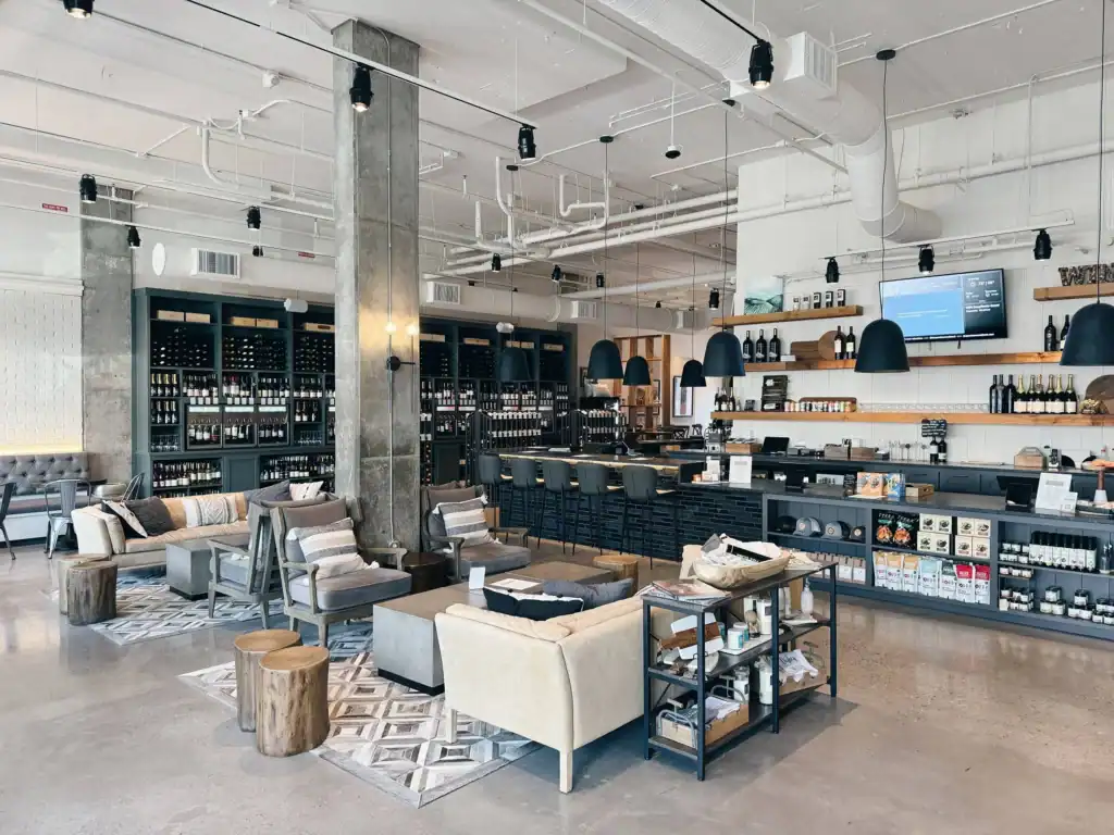 Modern café interior with lounge seating, wooden tables, shelves stocked with bottles, and a bar counter. The space features industrial design elements and neutral tones.