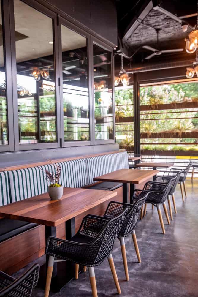 A modern indoor café seating area with wooden tables, black chairs, striped bench cushions, and a potted plant on the table near large windows.