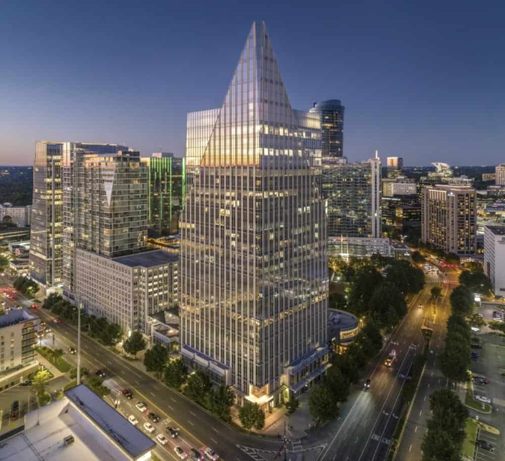 A tall, glass office building with a triangular top stands among other modern high-rises in a cityscape at dusk.