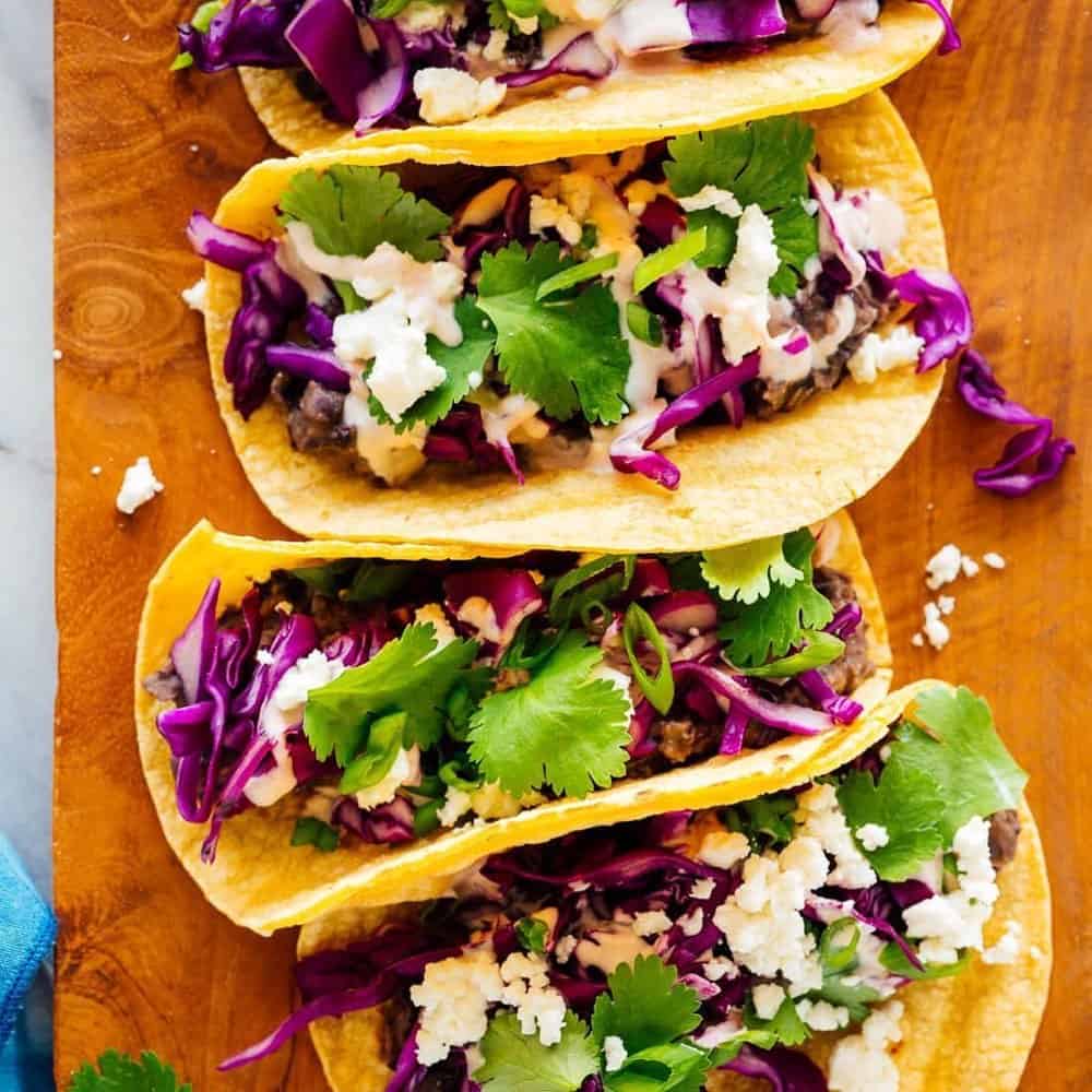 Three tacos on a wooden board, filled with black beans, shredded purple cabbage, cilantro, crumbled cheese, and drizzled with a creamy sauce.