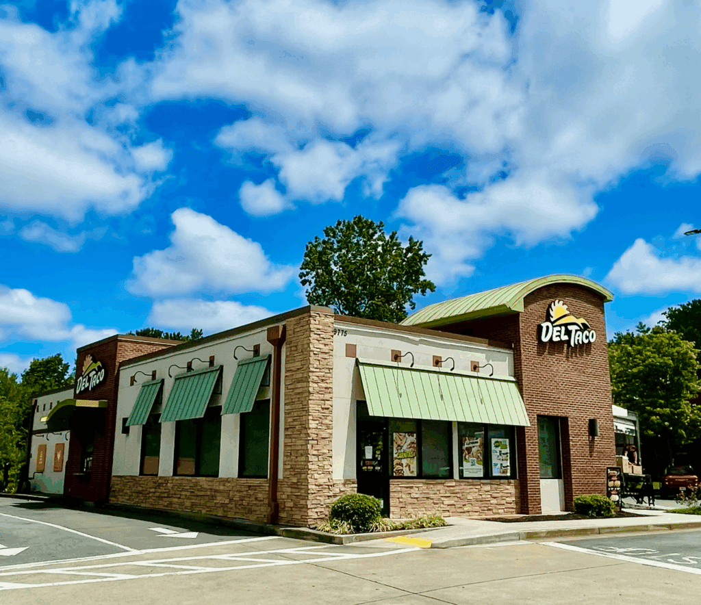 A Del Taco fast food restaurant with green awnings and a brick exterior, featuring a QSR drive thru, pictured on a sunny day with a blue sky and scattered clouds.